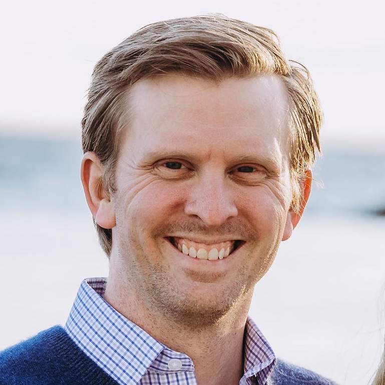 Close outdoor shot of Ben smiling broadly in a checkered blue collared shirt, with an ocean horizon in soft focus in the background.