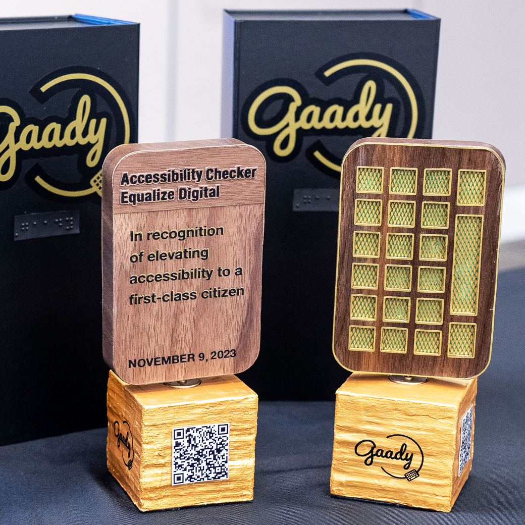2 of the 3 Gaadys trophies, one which reads 'In recognition of elevating accessibility to a first-class citizen' and the other displaying the back side of the trophy, a wood keyboard design with dark green, textured material for the keys. Both are on a gold cube base with the word 'Gaady' and a QR code.